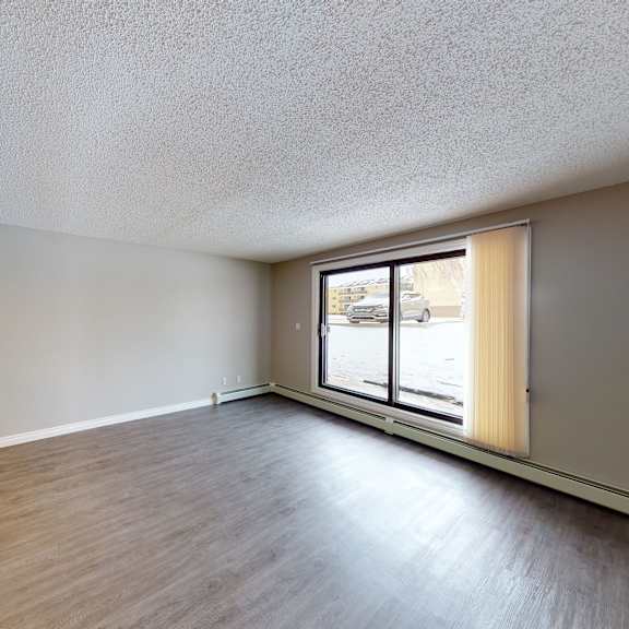 empty living room here at wyndham crossing apartment homes with gray walls and dark wood flooring, featuring a large window with vertical blinds. natural light creates a calm, open atmosphere.