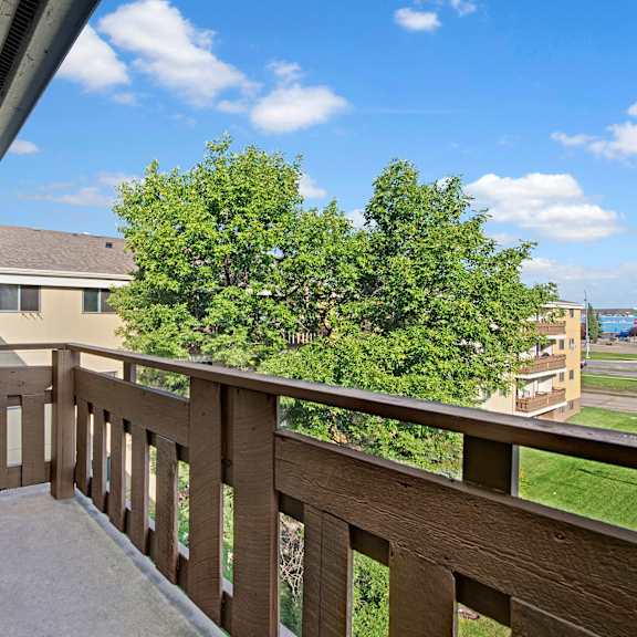 balcony view here at wyndham crossing apartment homes with brown railing overlooking a tree and distant buildings under a bright blue sky with scattered clouds, conveying a peaceful, open-air atmosphere.