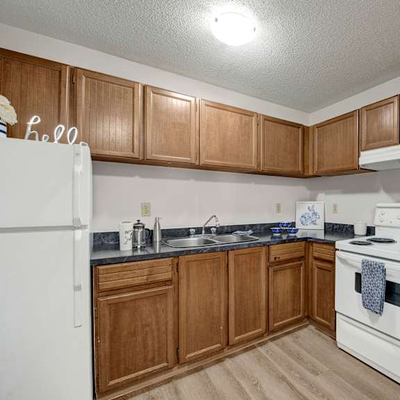 compact kitchen here at wyndham crossing apartment homes with brown cabinets, white appliances, and a black countertop. includes a fridge, oven, and sink. bright, clean, and organized space.