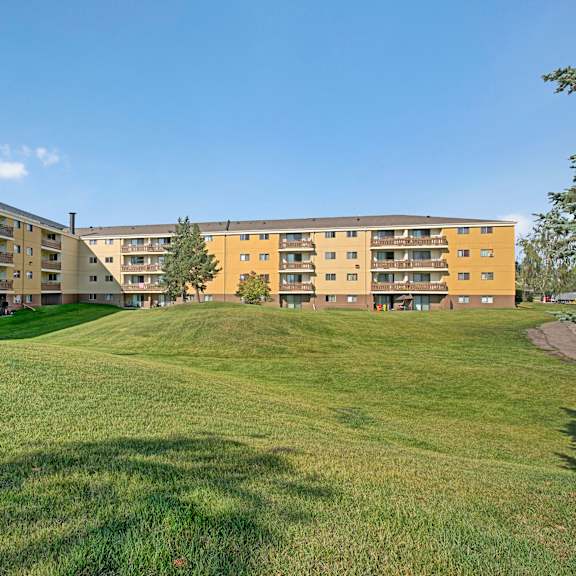 wyndham crossing apartment homes complex with yellow and brown buildings, surrounded by lush green grass and trees under a blue sky. peaceful, sunny atmosphere.
