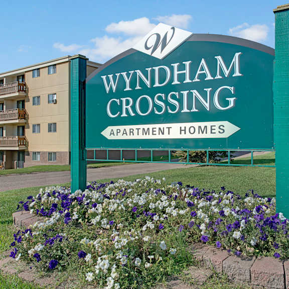 sign reading "wyndham crossing apartment homes" stands on green lawn, bordered by purple and white flowers. apartment building and trees in background.
