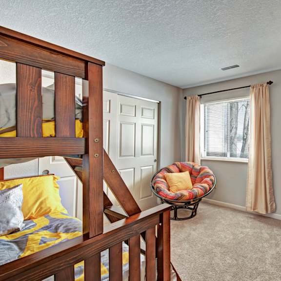 a cozy bedroom here at river’s cove featuring a wooden bunk bed with gray and yellow bedding, soft carpet, and a dark dresser with lamp, creating a warm, comfortable, and inviting space.