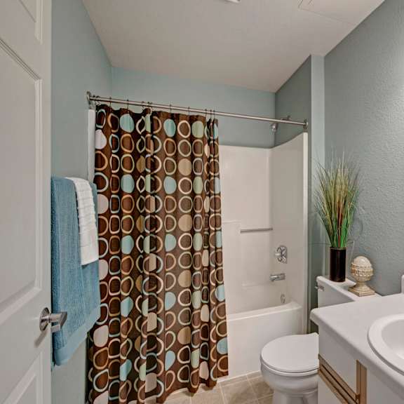 a clean bathroom here at river’s cove featuring a bright vanity, large mirror, patterned shower curtain, and blue accent towels, creating a fresh, inviting, and well-lit space.