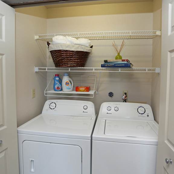 a convenient laundry room here at river’s cove featuring full-size washer and dryer, open shelving, and organized storage, creating a clean, practical, and functional space.