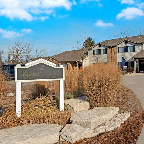 a welcoming exterior here at river’s cove featuring the community entrance sign, attached garages, and landscaped surroundings, creating a charming, well-kept, and inviting first impression.