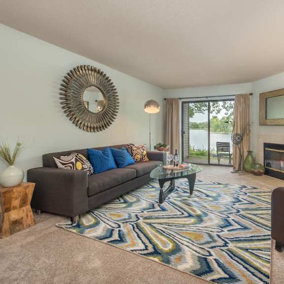 a bright living room here at river’s cove with a gray sofa and blue accent pillows, patterned blue rug, and cozy fireplace. large sliding doors open to a scenic lake view, creating a relaxing, welcoming atmosphere.