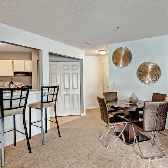 a cozy dining area here at river’s cove with a round table, brown upholstered chairs, and gold wall accents. the breakfast bar with black stools adds a welcoming space for casual dining and entertaining.