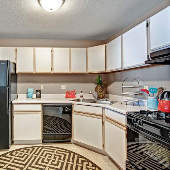 a bright kitchen here at river’s cove featuring white cabinetry, black appliances, and a welcoming layout. a patterned rug and small décor accents add charm, creating a clean, inviting space for everyday cooking and dining.