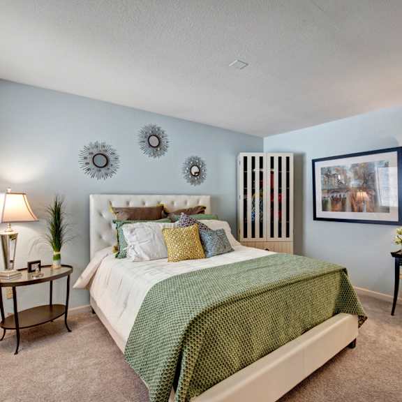 a cozy bedroom here at river’s cove featuring a plush bed with green bedding, soft carpet, and calming blue walls. matching lamps and tasteful décor create a warm, inviting retreat perfect for relaxing.