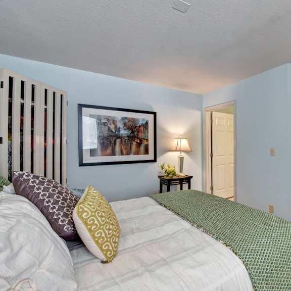 a bright bedroom here at river’s cove featuring a plush bed with green bedding, soft carpet, and calming blue walls. a side table with lamp and framed artwork adds a warm, welcoming touch.