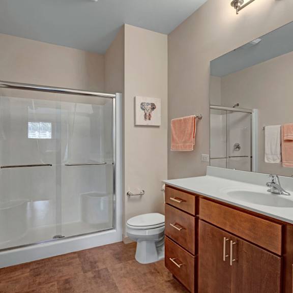 a clean, modern bathroom here at vue at pinnacle park with a wide vanity and mirror, wood cabinets, bright lighting, a full shower with sliding glass doors, and warm-toned flooring for a polished, comfortable feel.