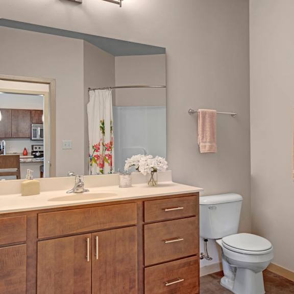 a bright bathroom here at vue at pinnacle park with a wide vanity and mirror, wood cabinets, white countertops, a tub-shower combo with curtain, and warm flooring for a clean, comfortable, and inviting feel.