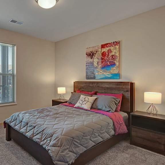 a bright bedroom here at vue at pinnacle park with soft carpet, a large window with blinds and neighborhood views, a wood bed frame with gray and pink bedding, and matching nightstands with warm bedside lighting.