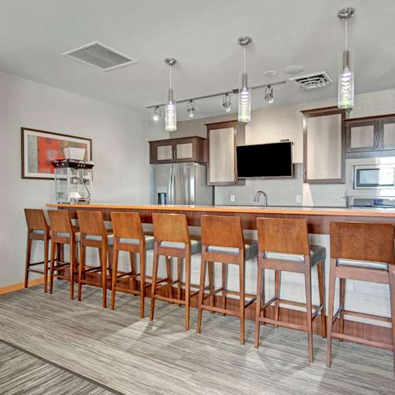 a modern clubhouse kitchen here at vue at pinnacle park with a long bar counter, multiple bar stools, stainless steel appliances, pendant lighting, and a built-in tv for convenient dining and social gatherings.