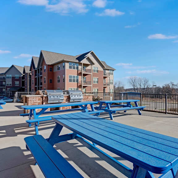 a sunny outdoor grilling area here at vue at pinnacle park with built-in barbecue grills, bright blue picnic tables, open patio space, and wide scenic views for relaxed outdoor dining and gatherings.