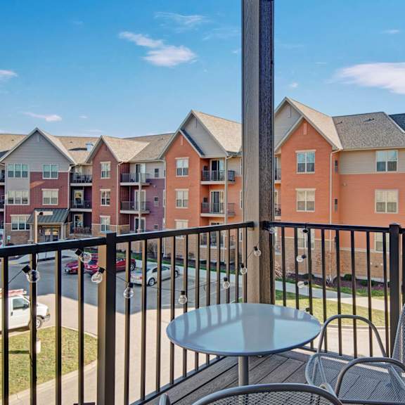 a private apartment balcony here at vue at pinnacle park with a small patio table and chairs, railing with string lights, and open views of nearby buildings and landscaped grounds.