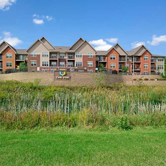 a scenic exterior view here at vue at pinnacle park with multi-story apartment buildings, private balconies, landscaped grounds, and a pond with natural greenery creating a peaceful community setting.