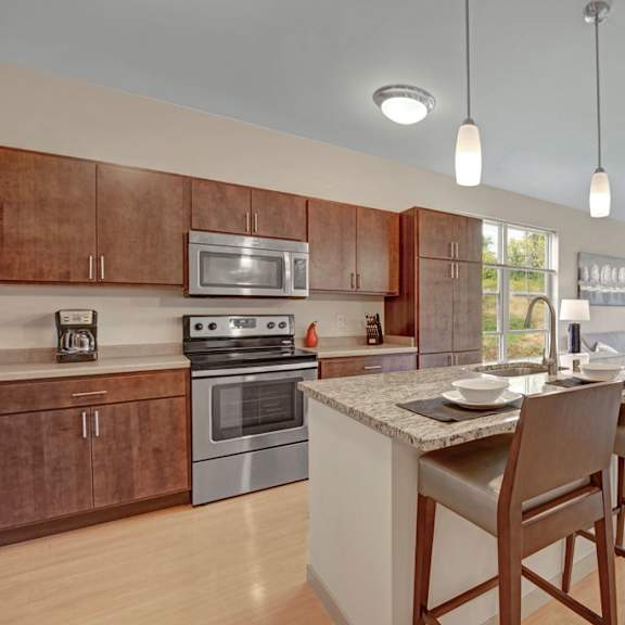 a modern open kitchen and living area here at vue at pinnacle park with warm wood cabinets, stainless steel appliances, granite island with bar seating, pendant lighting, and large windows bringing in bright natural light.