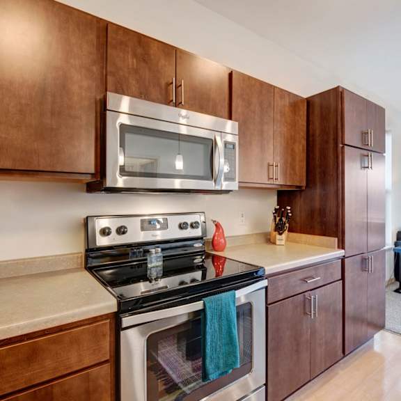 a modern kitchen here at vue at pinnacle park with rich wood cabinets, stainless steel stove and microwave, smooth countertops, and generous storage, opening toward a bright, comfortable living area with soft carpet and natural light.