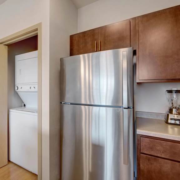 a convenient in-unit laundry area here at vue at pinnacle park with a stacked washer and dryer behind a closet door, next to a stainless steel refrigerator and warm wood kitchen cabinets in a clean, modern layout.