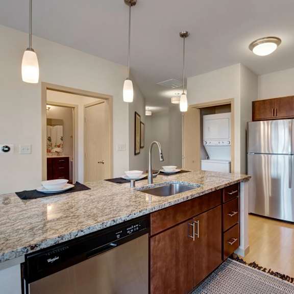 a modern kitchen here at vue at pinnacle park with granite countertops, a large island with sink and bar seating, stainless steel appliances, pendant lighting, and an in-unit stacked washer and dryer nearby for added convenience.