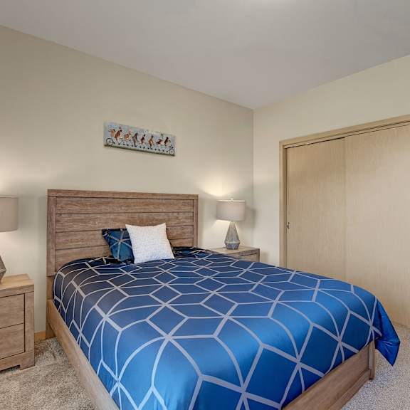 a comfortable bedroom here at vue at pinnacle park with soft carpet, a wood bed frame, blue patterned bedding, matching nightstands with lamps, and a wide sliding-door closet in a clean, cozy layout.