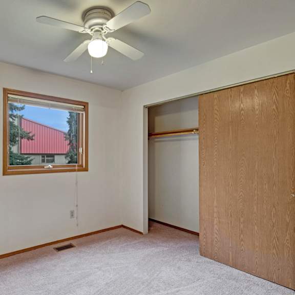 plain bedroom here at wildwood estates apartments with beige carpeting and white walls, featuring a ceiling fan. a window shows a view of a tree and red-roofed building. open closet with wooden sliding door.
