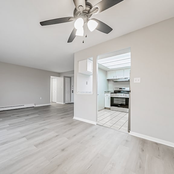 a spacious room with a ceiling fan and light, hardwood floors, and a kitchen visible through an open door.