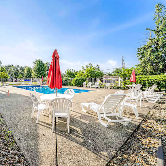 a pool area with chairs and umbrellas is surrounded by a gravel path and greenery.