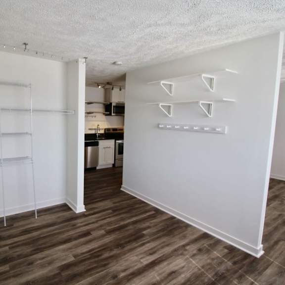 a room with white walls and wooden flooring with a kitchen in the background.