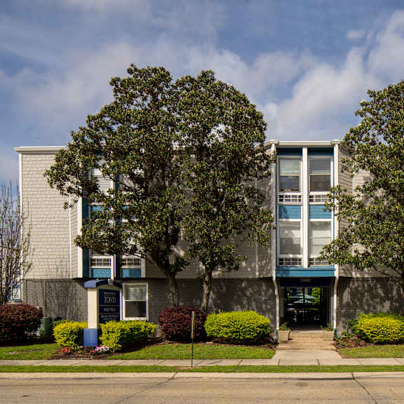 a building with a grey facade and a blue awning is surrounded by green bushes and trees.