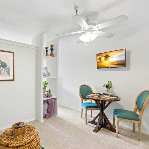 a room with a ceiling fan, a painting on the wall, a small table with chairs and a basket on the floor.