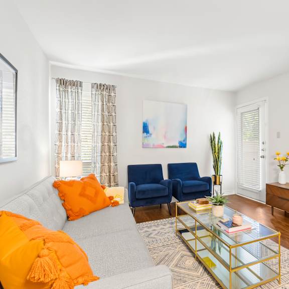 a living room with a grey couch, a blue chair, a glass coffee table, and a painting on the wall.