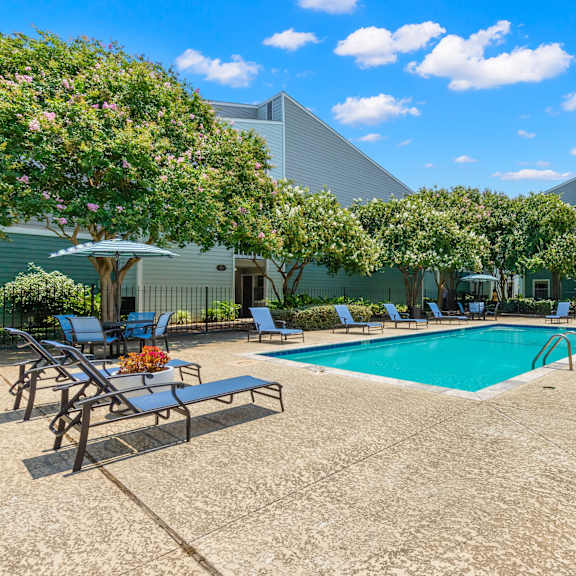 a pool surrounded by chairs and trees with a building in the background.
