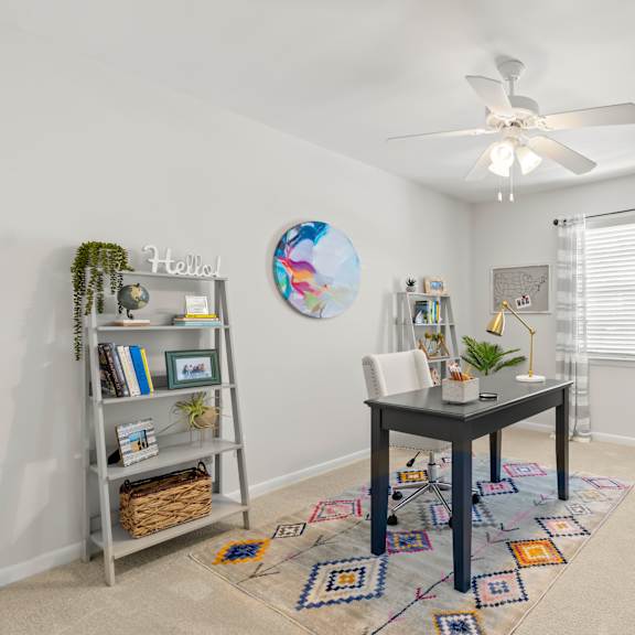 a room with a round wall clock, a bookshelf, a table, a chair, and a rug.