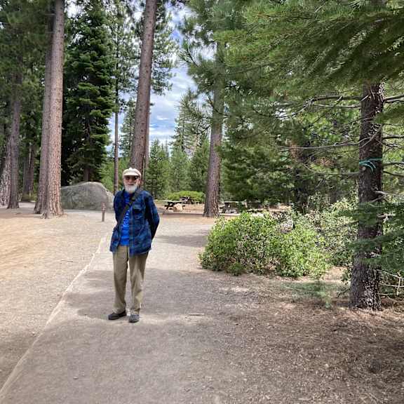 a man in a blue jacket and khaki pants stands on a dirt path in a forest.