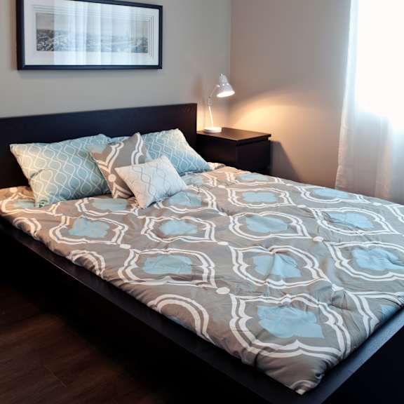 a bed with a grey and white patterned comforter and two blue patterned pillows sits in a room with a framed picture on the wall and a lamp on a side table.