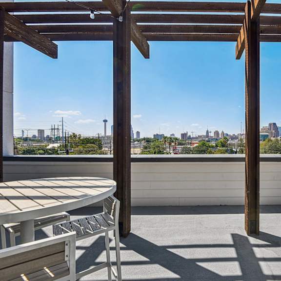 a wooden pergola with a table and chairs is set up on a rooftop with a cityscape in the background.