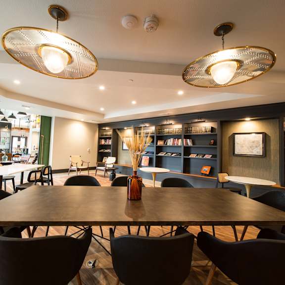 a large wooden table surrounded by black chairs in a room with a bookshelf and a chalkboard.