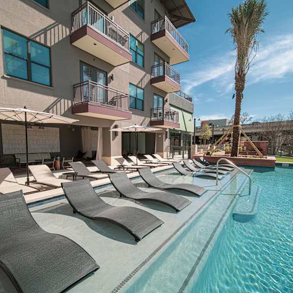a pool with lounge chairs and umbrellas in front of a building with balconies.
