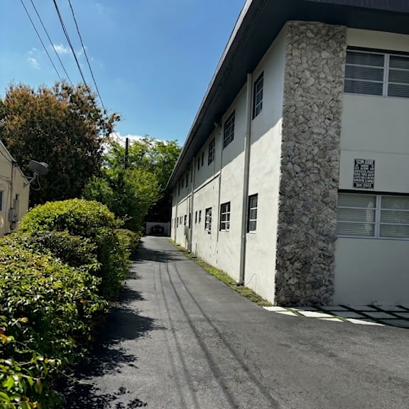a long, narrow road with a white building on one side and a hedge on the other.