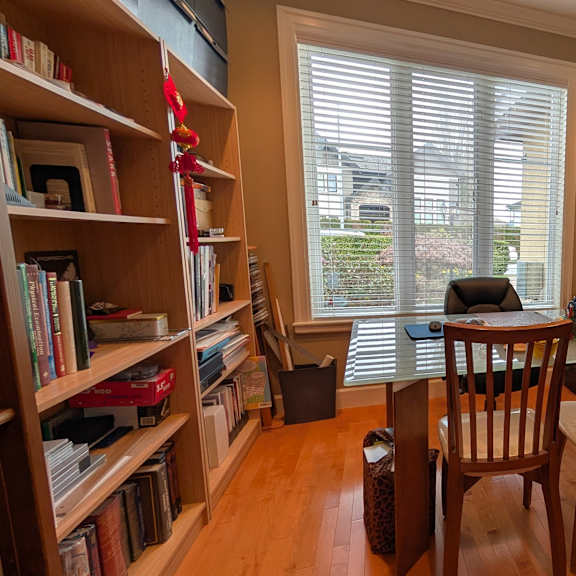 a room with a bookshelf, a chair, and a window with blinds.