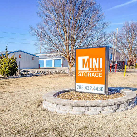 a mini storage sign with a phone number is in the foreground of a grassy area with a building in the background.