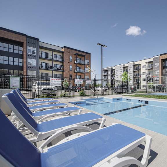 a pool surrounded by blue lounge chairs and apartment buildings.