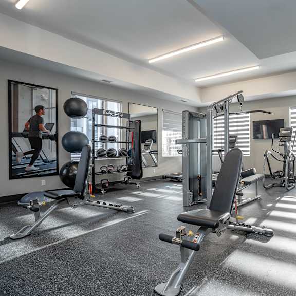 a well-equipped gym with various exercise machines and a person working out.at vibe apartments at arbour lake, calgary