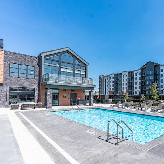 a swimming pool in a sunny day surrounded by chairs and buildings.