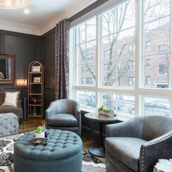 a living room with a grey ottoman, grey armchair, and a grey couch with a white throw.