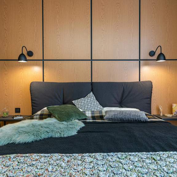 a bed with a floral comforter and a green pillow is in the foreground of a room with two wall lamps.
