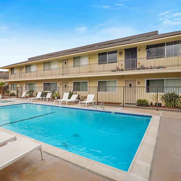 a swimming pool surrounded by sun loungers in front of apartment buildings.