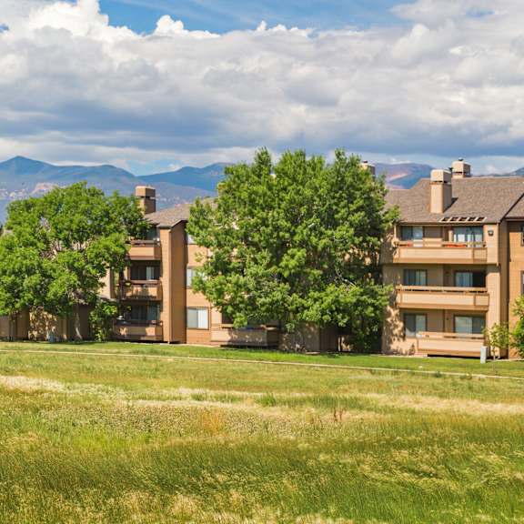 a grassy field in front of apartment buildings with trees and mountains in the background.